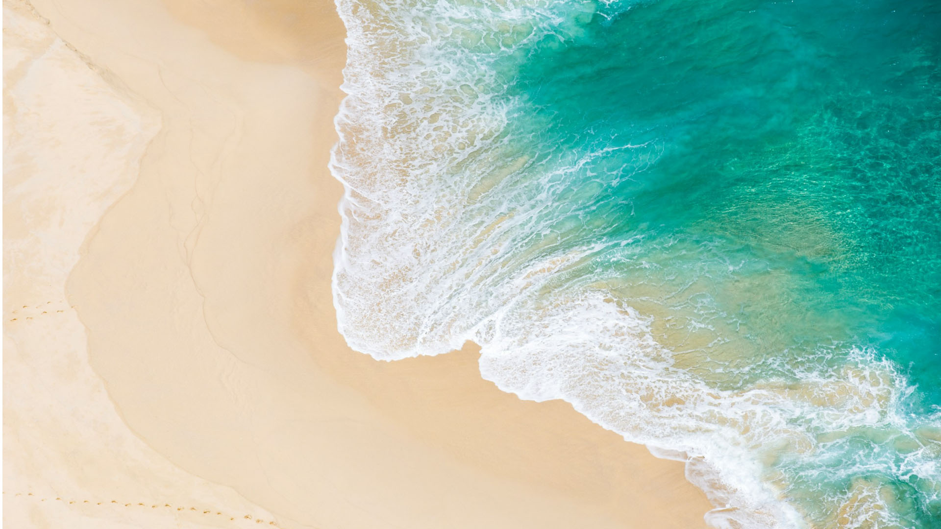 Top-down view of turquoise surf foaming onto a smooth, pale sandy beach.